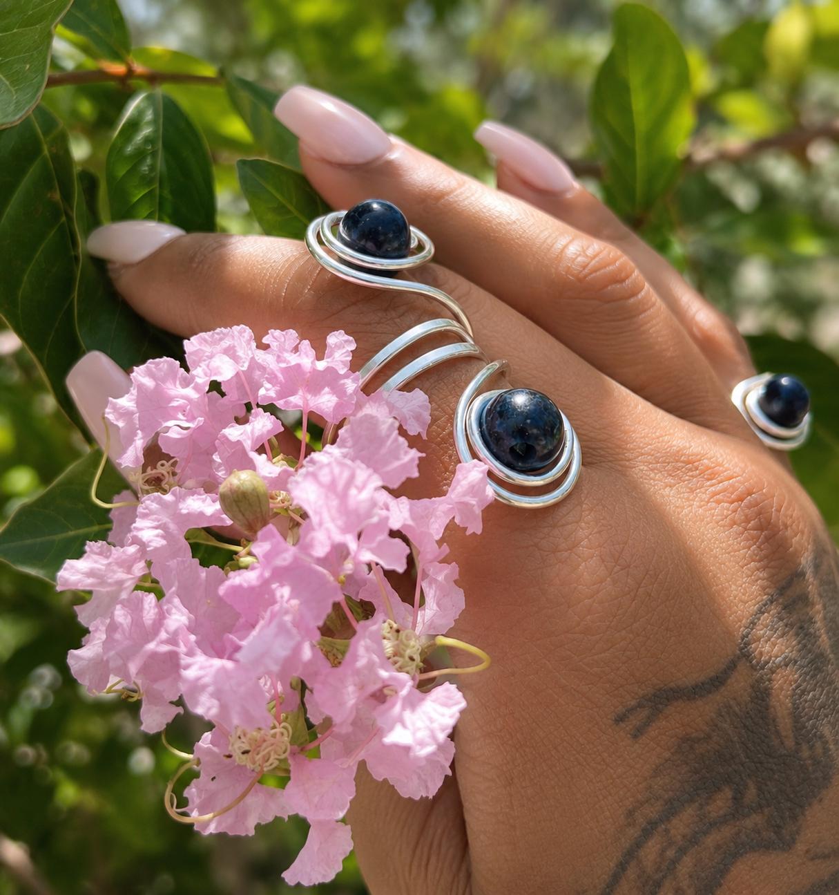 The Twin Moons Ring worn on the hand, two deep blue tiger eye stones cradled in flowing silver spirals, held against pink blossom.